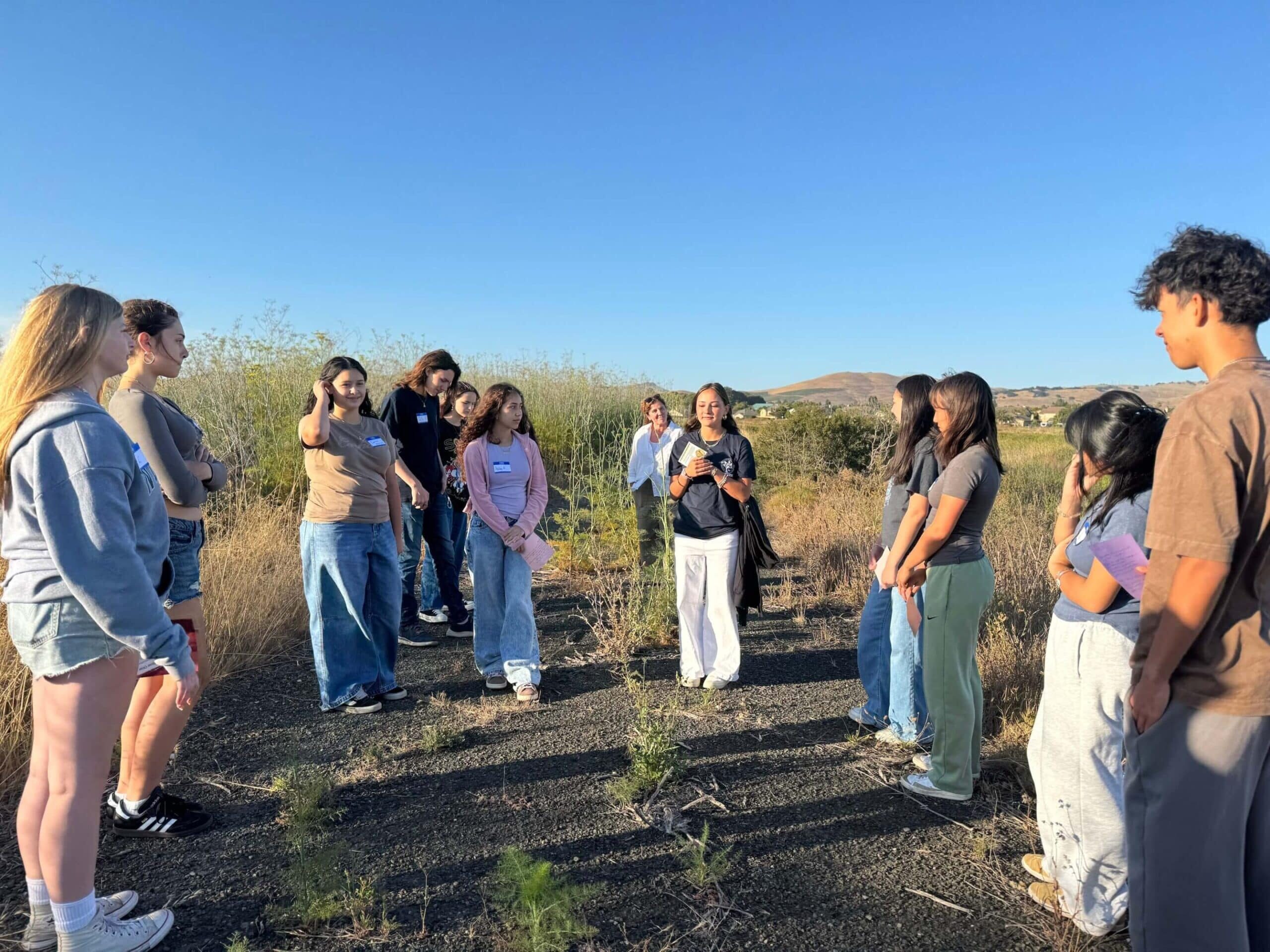 Teen WILD Volunteers teaching back on a nature walk. teens standing in a circle with a blue sky behind them